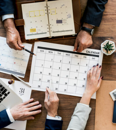 The image shows a business meeting with three professionals gathered around a table, reviewing multiple calendars and digital devices. They are discussing schedules or project timelines, indicated by one individual pointing to a specific date on a large printed calendar, surrounded by a laptop, mobile phones, a notebook, and various planning materials.