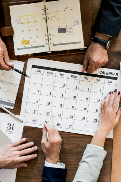 The image shows a business meeting with three professionals gathered around a table, reviewing multiple calendars and digital devices. They are discussing schedules or project timelines, indicated by one individual pointing to a specific date on a large printed calendar, surrounded by a laptop, mobile phones, a notebook, and various planning materials.